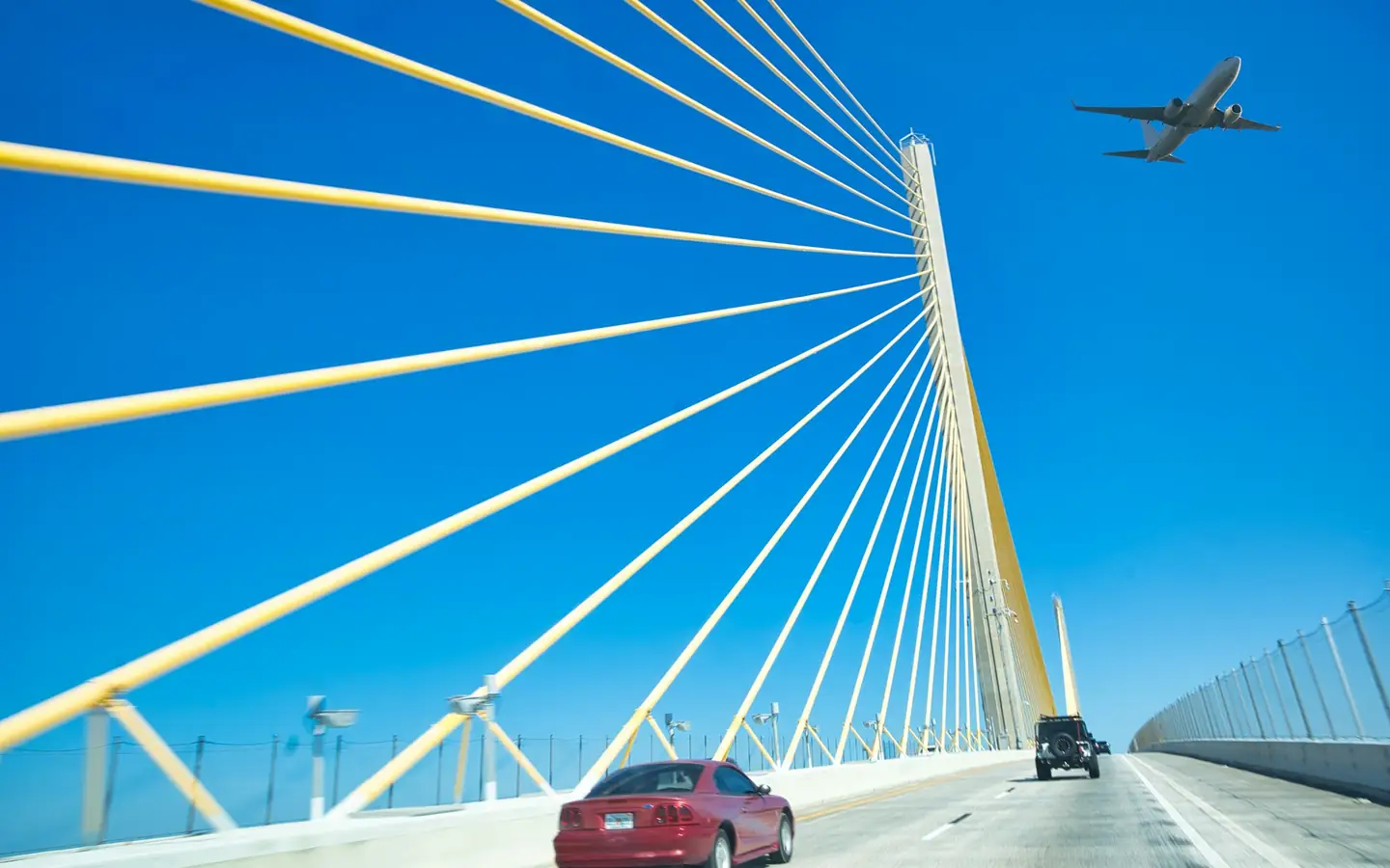 A red car crossing a cable-stayed bridge as a plane flies overhead